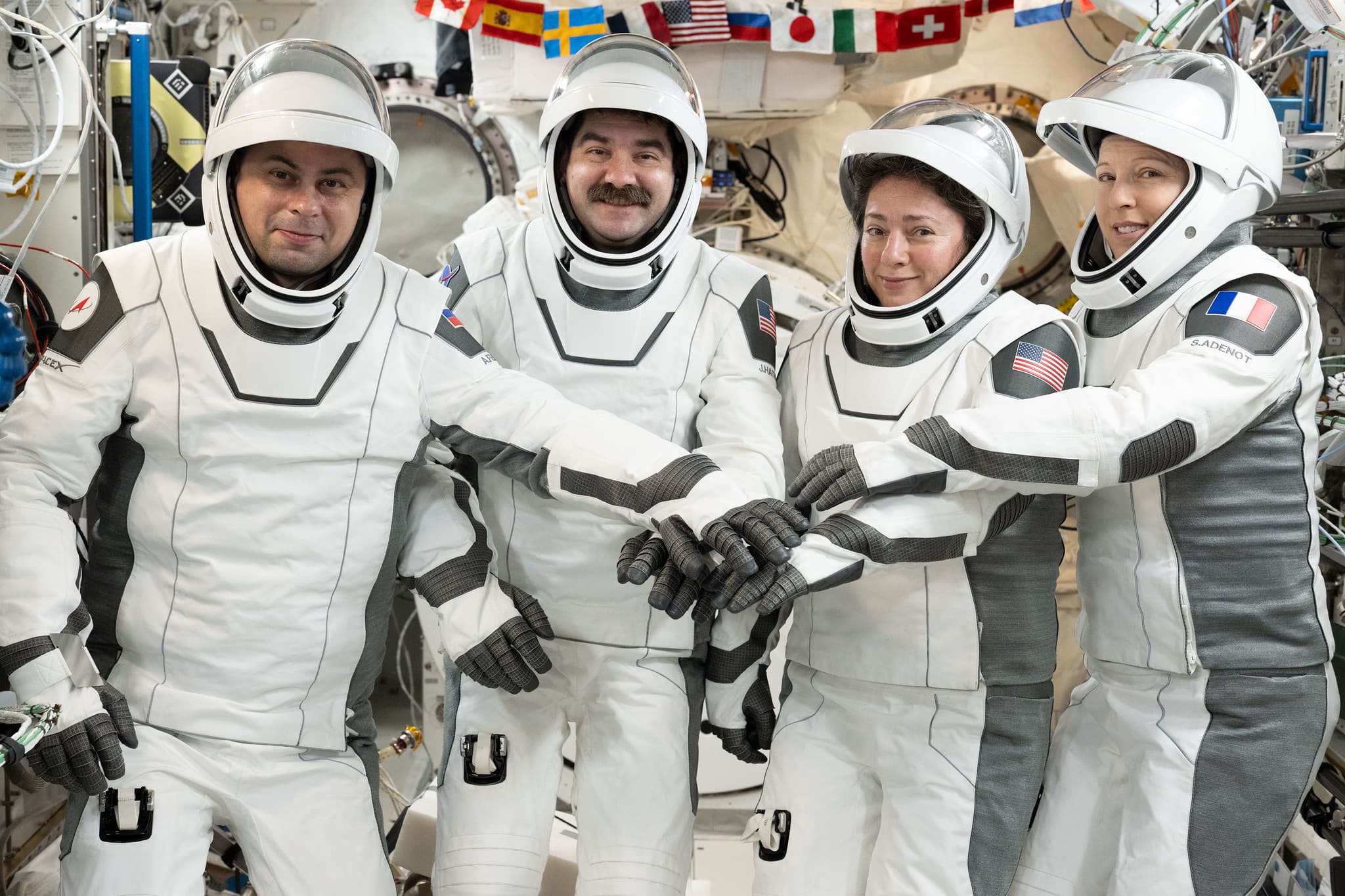 Four Crew-12 astronauts in white pressure suits join their gloved hands for a group portrait