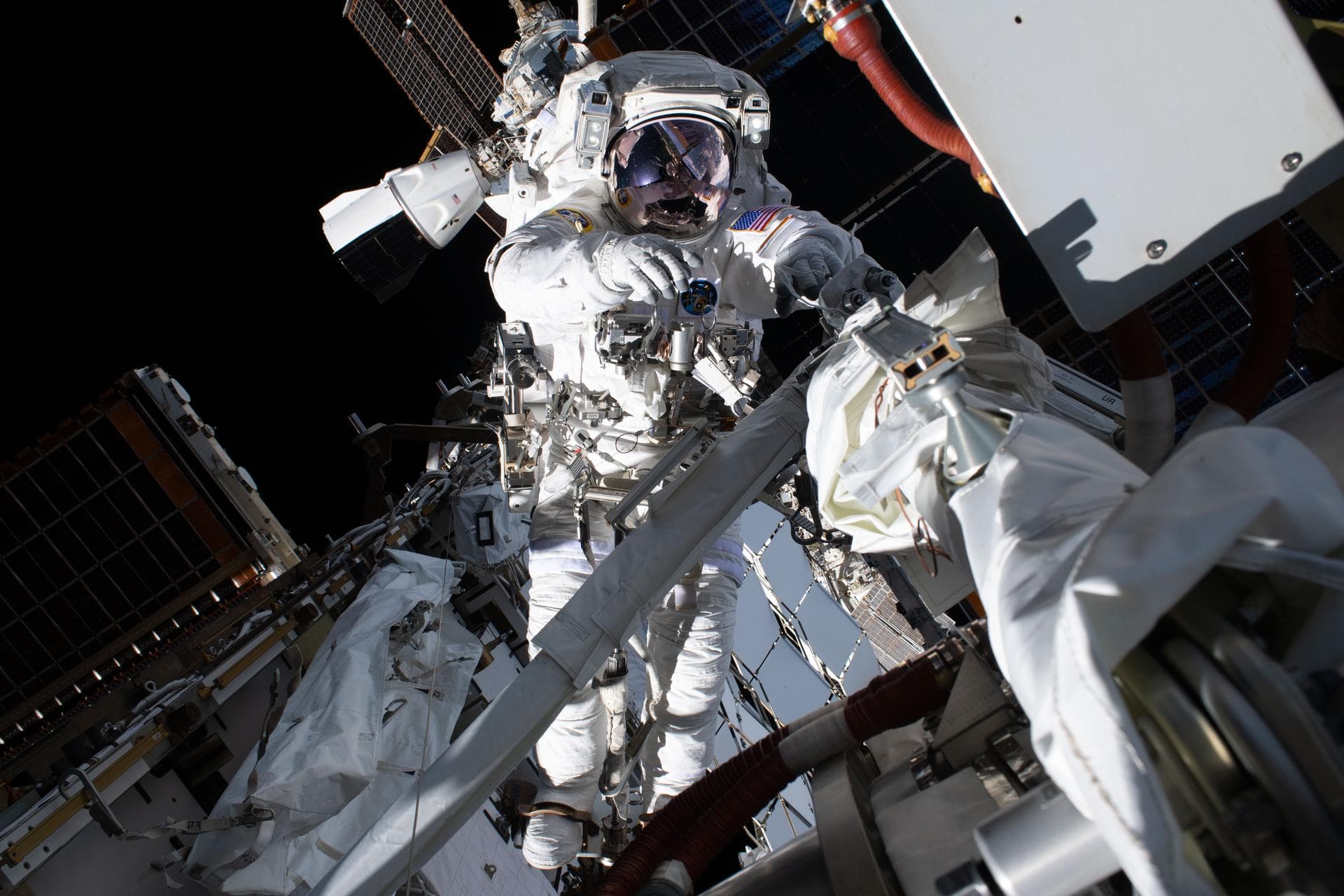 Anil Menon in a white spacesuit works among equipment and handrails outside the International Space Station