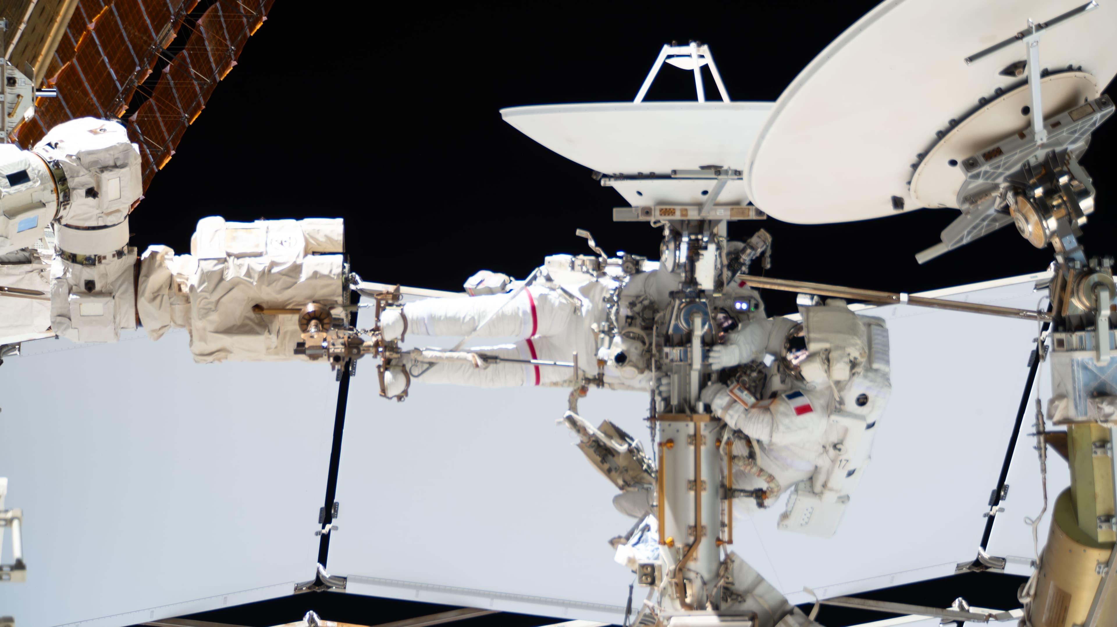 Two spacesuited astronauts work near a white dish antenna on the exterior of the International Space Station