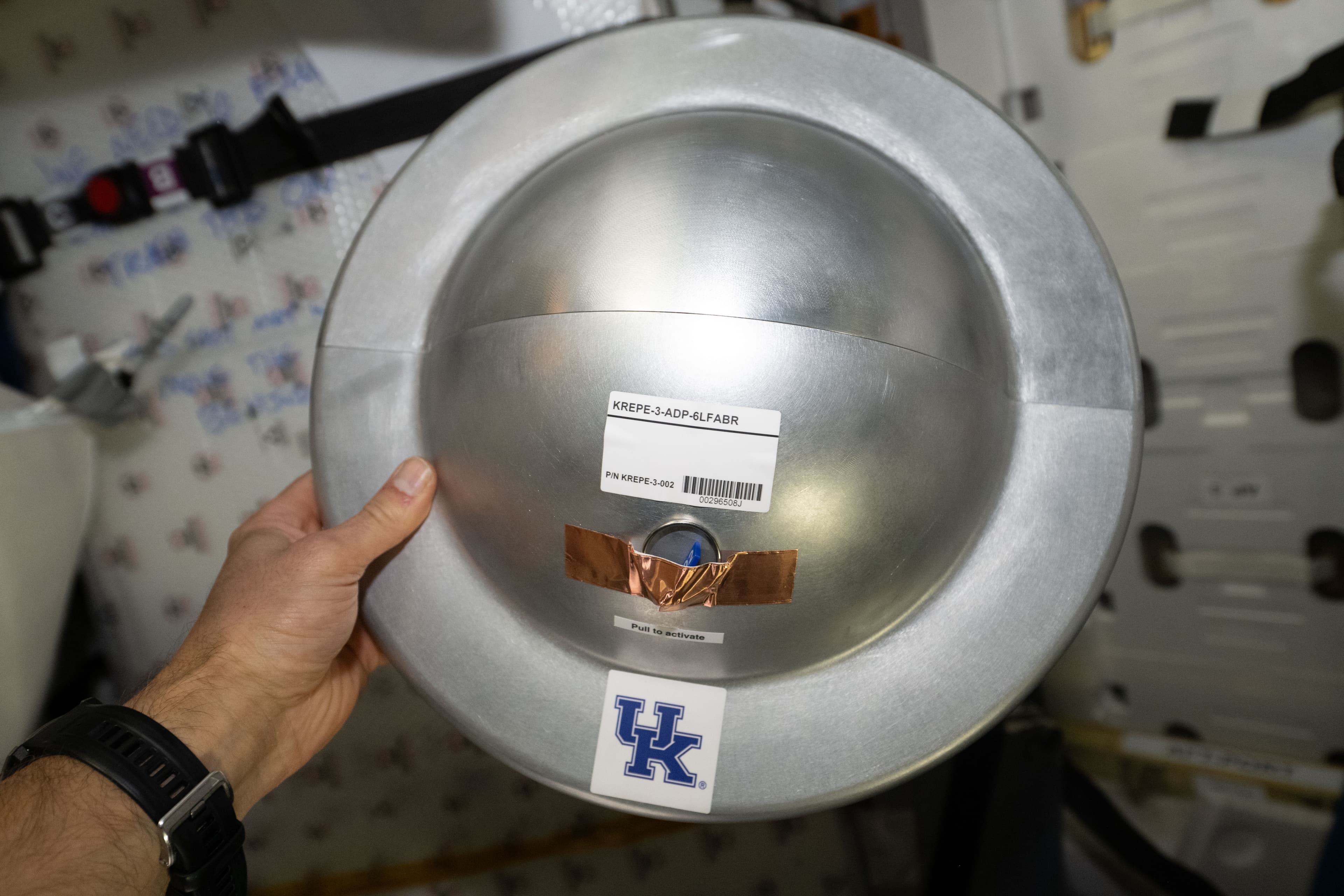 A hand holds a metallic experiment capsule inside Cygnus, with equipment labels and a University of Kentucky mark visible