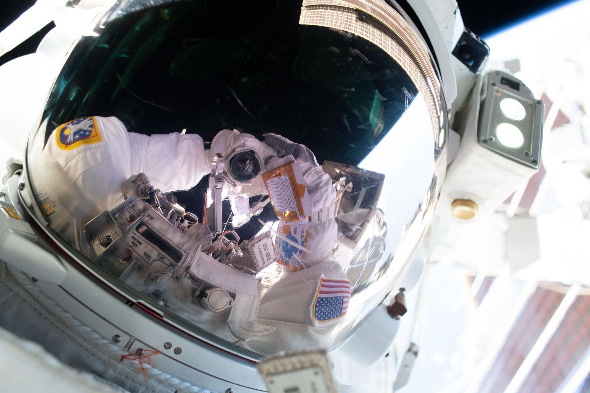 Anil Menon’s spacesuit and camera reflected in a helmet visor outside the space station