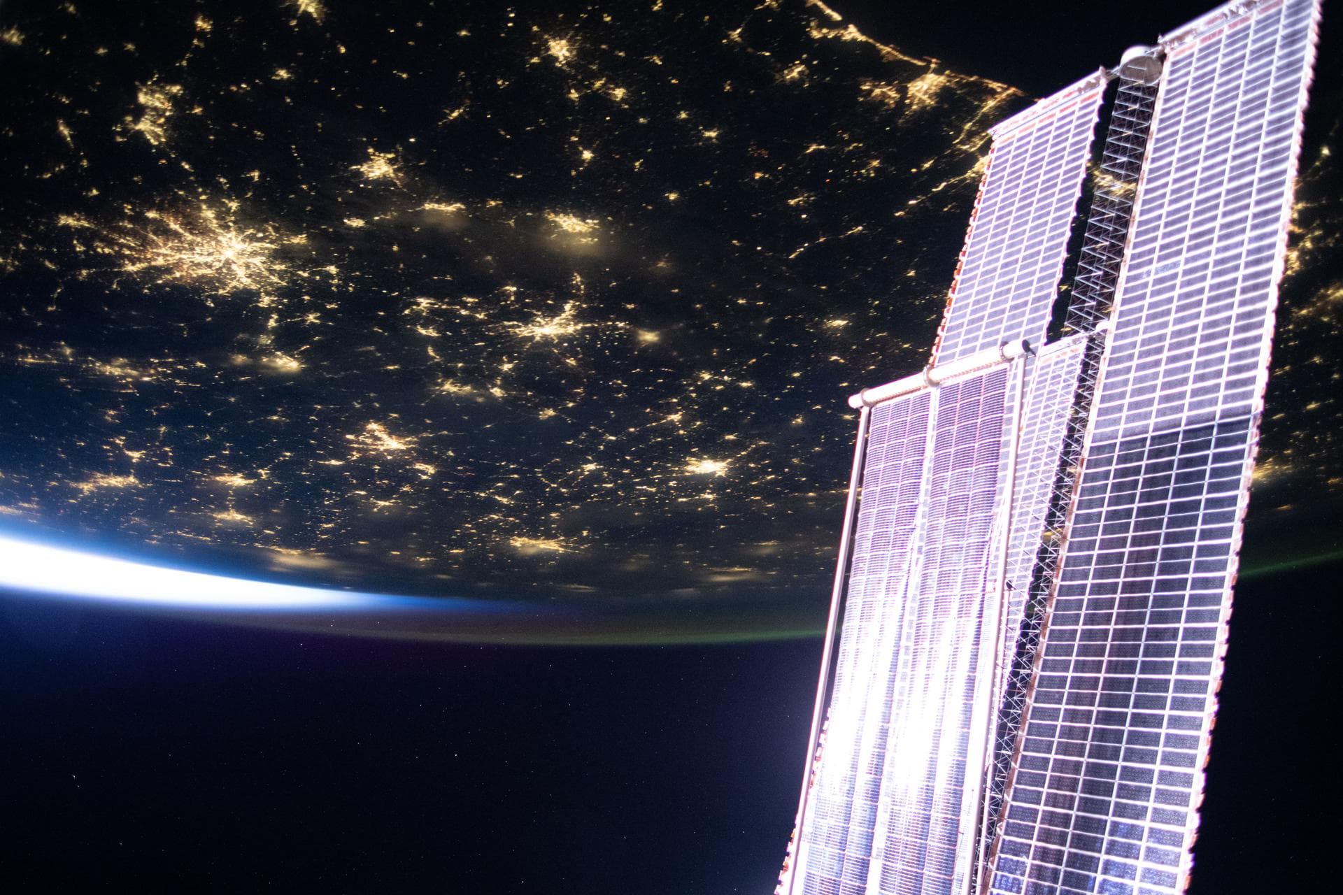 Large station solar panels and a smaller roll-out array above city lights and Earth’s bright horizon