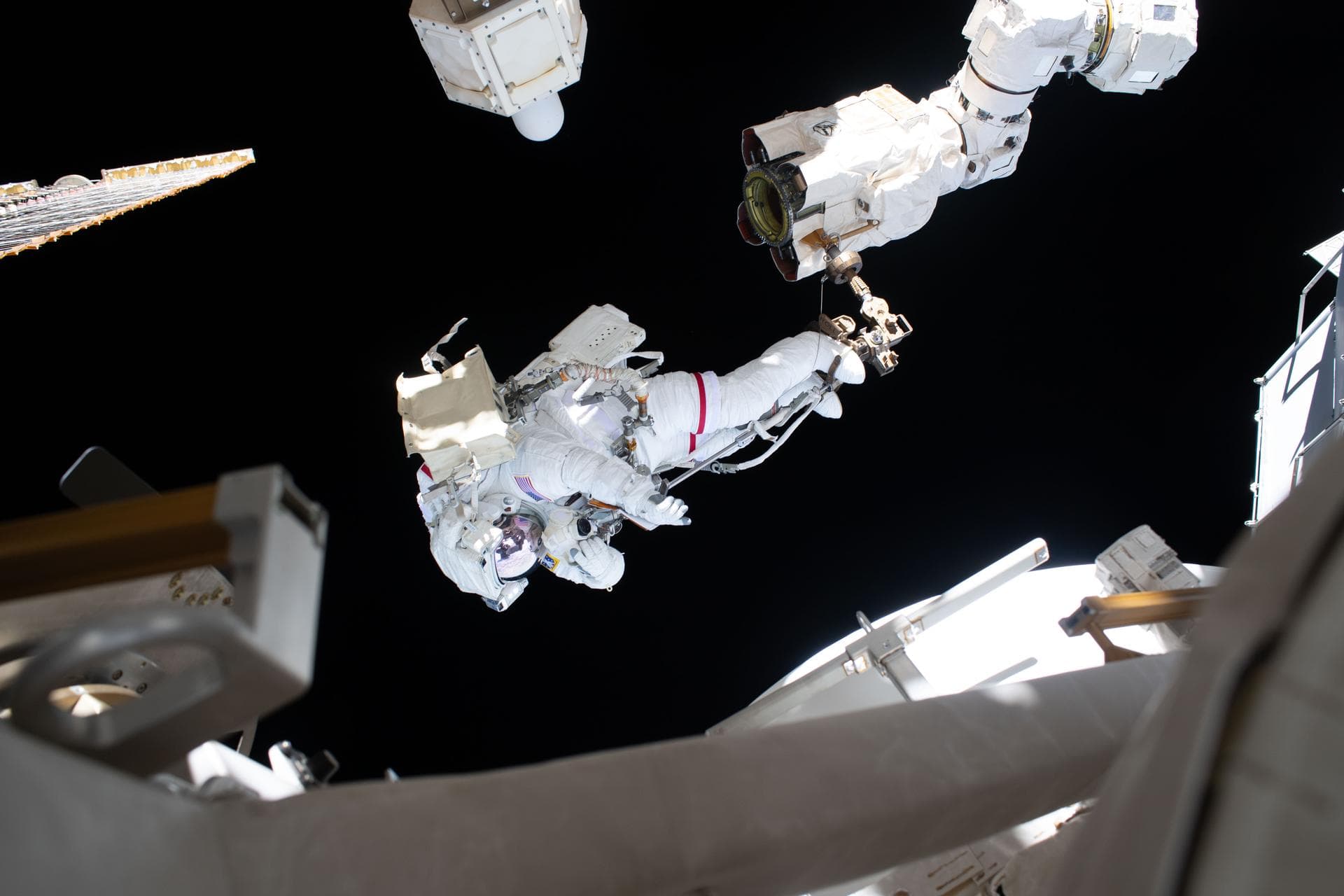 Anil Menon in a white spacesuit reaches toward Canadarm2 outside the International Space Station