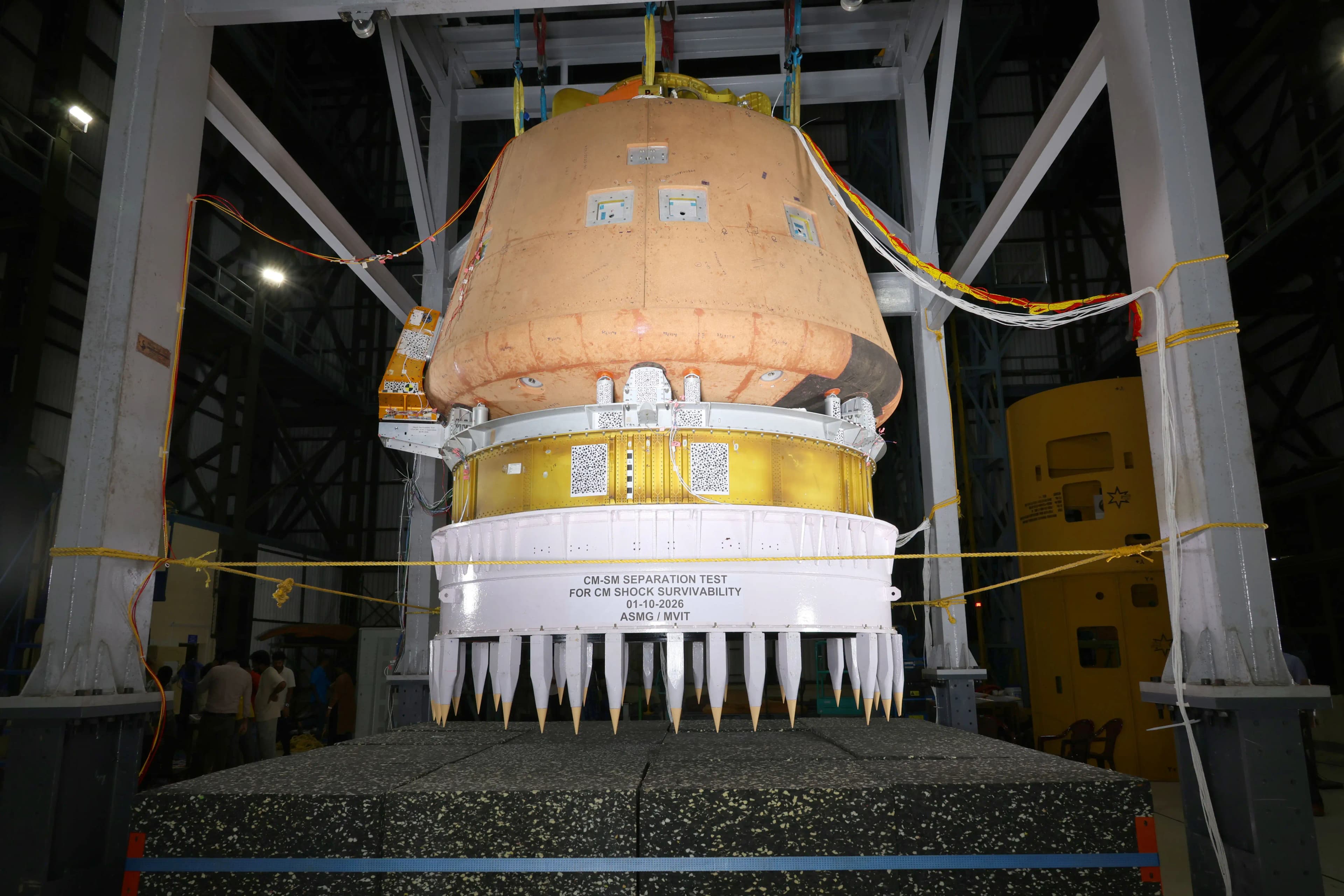 A capsule-shaped crew-module test assembly suspended above a cylindrical service-module rig inside a steel frame