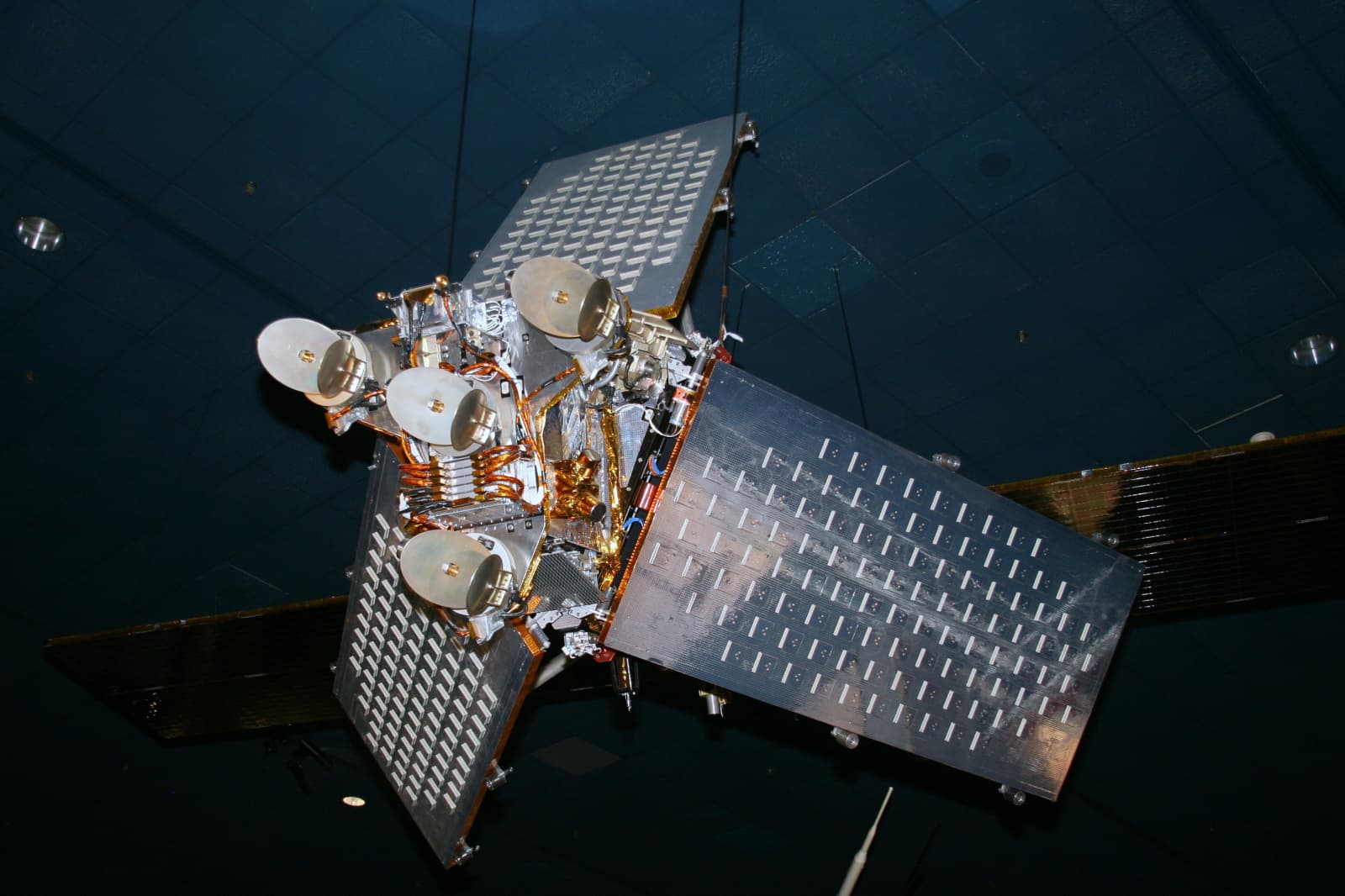 A silver Iridium satellite replica with three large antenna panels suspended indoors
