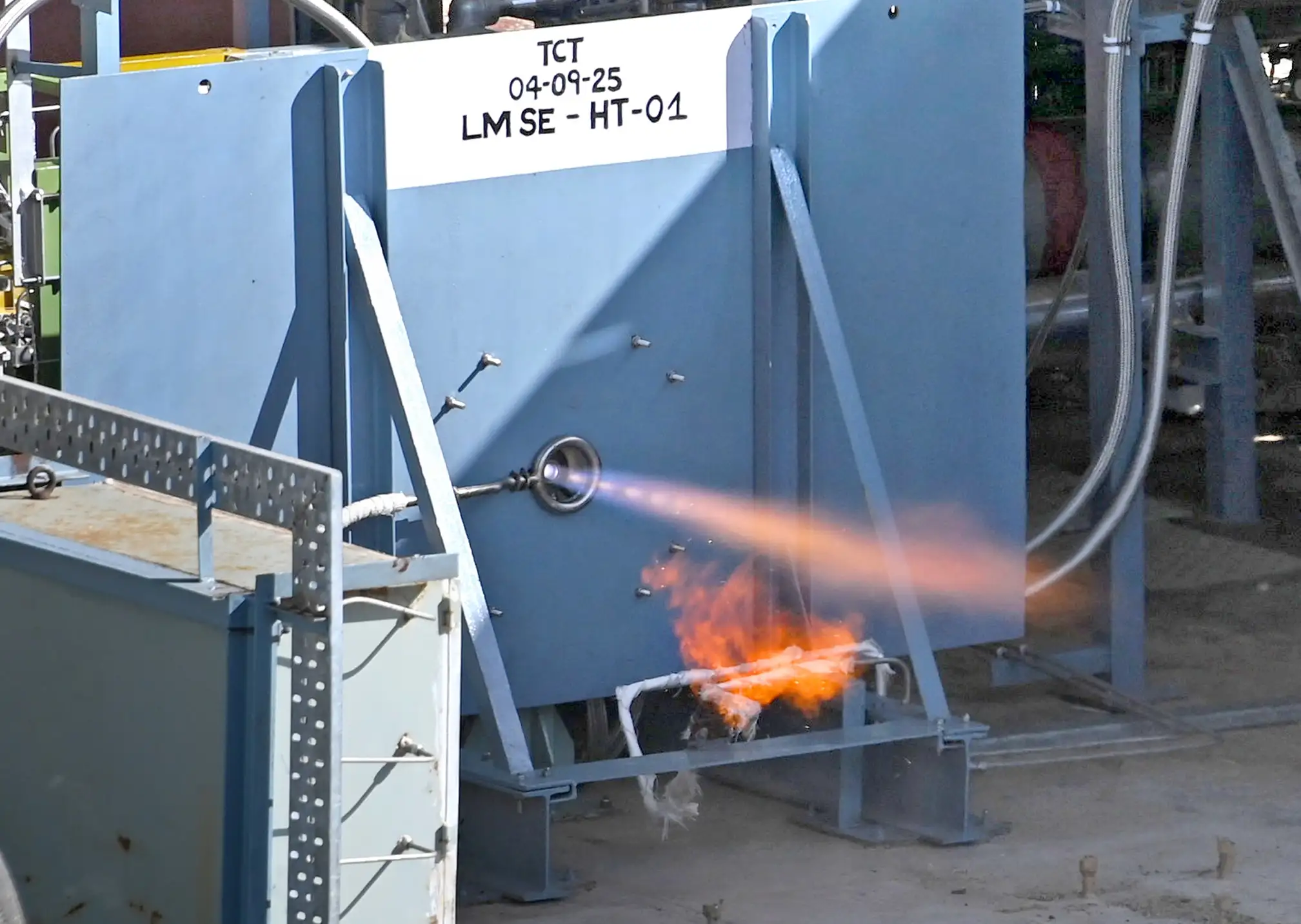 A small thrust chamber emits a narrow orange exhaust plume from a blue metal test stand