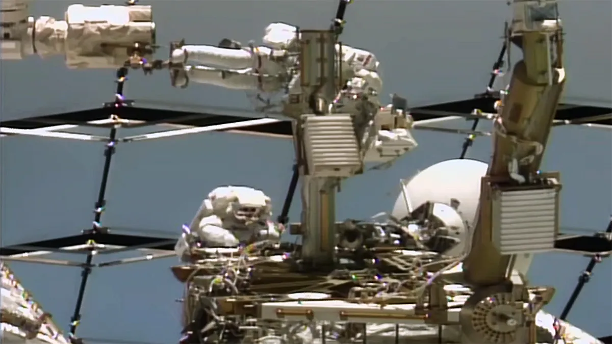 Two spacesuited astronauts work beside a white dish antenna on the exterior of the International Space Station