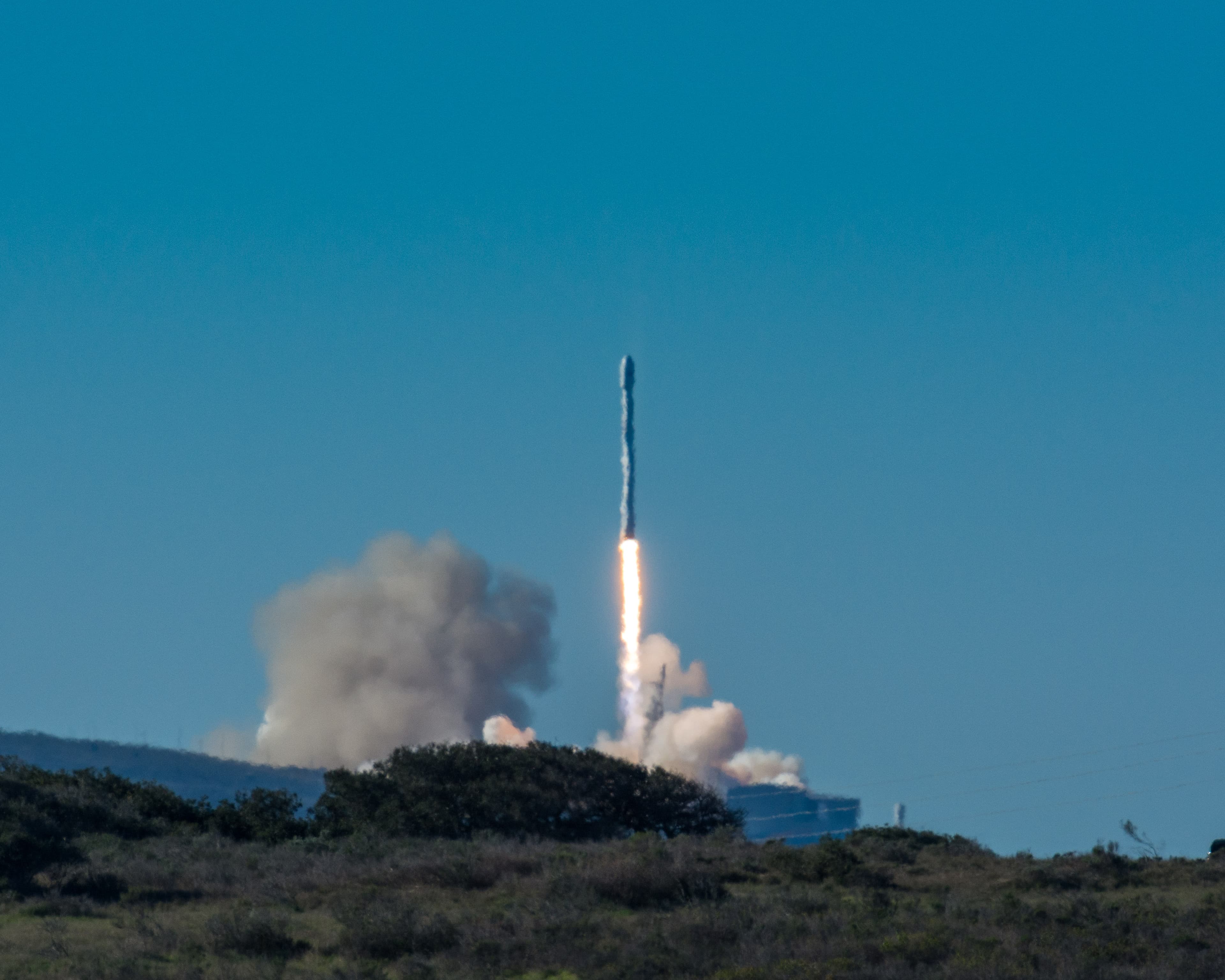 A Falcon 9 rocket climbs above scrubland with a bright exhaust plume and smoke near the launch pad