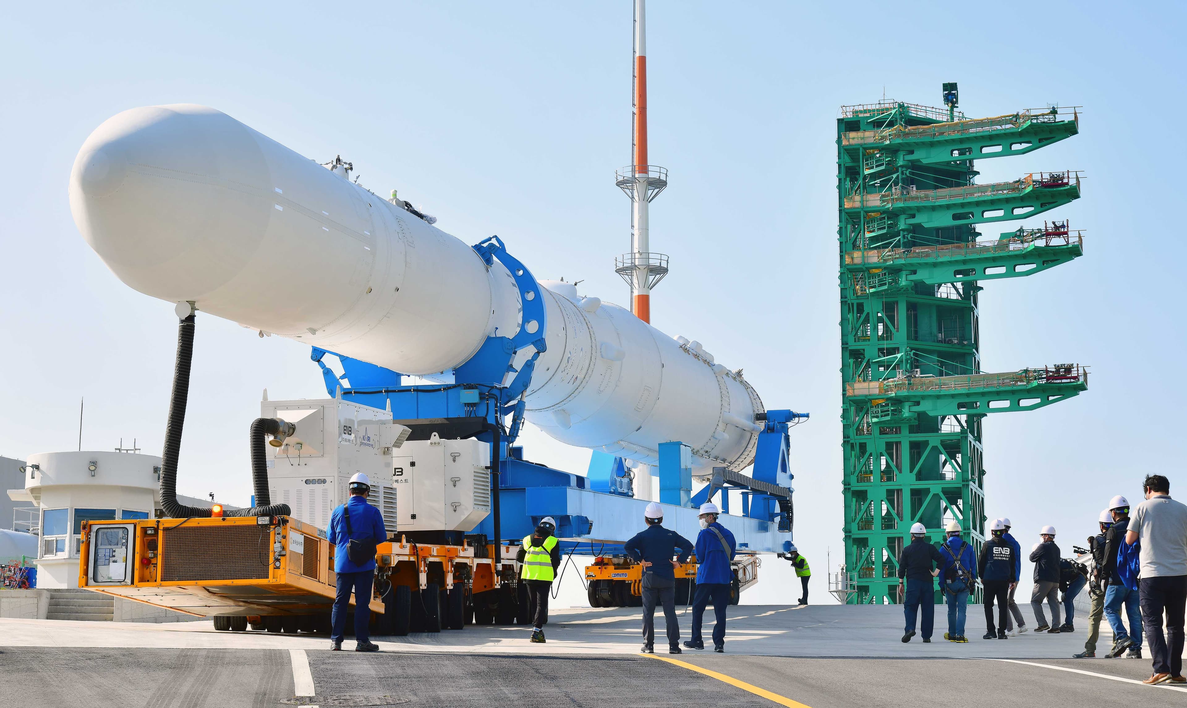 White Nuri rocket carried horizontally on a blue transporter beside a green launch tower, with workers on the pad