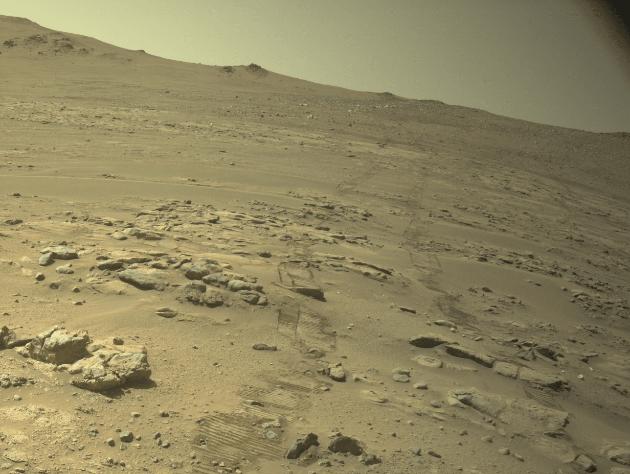Rover wheel tracks cross a sandy, rock-strewn Martian slope beneath a low ridge
