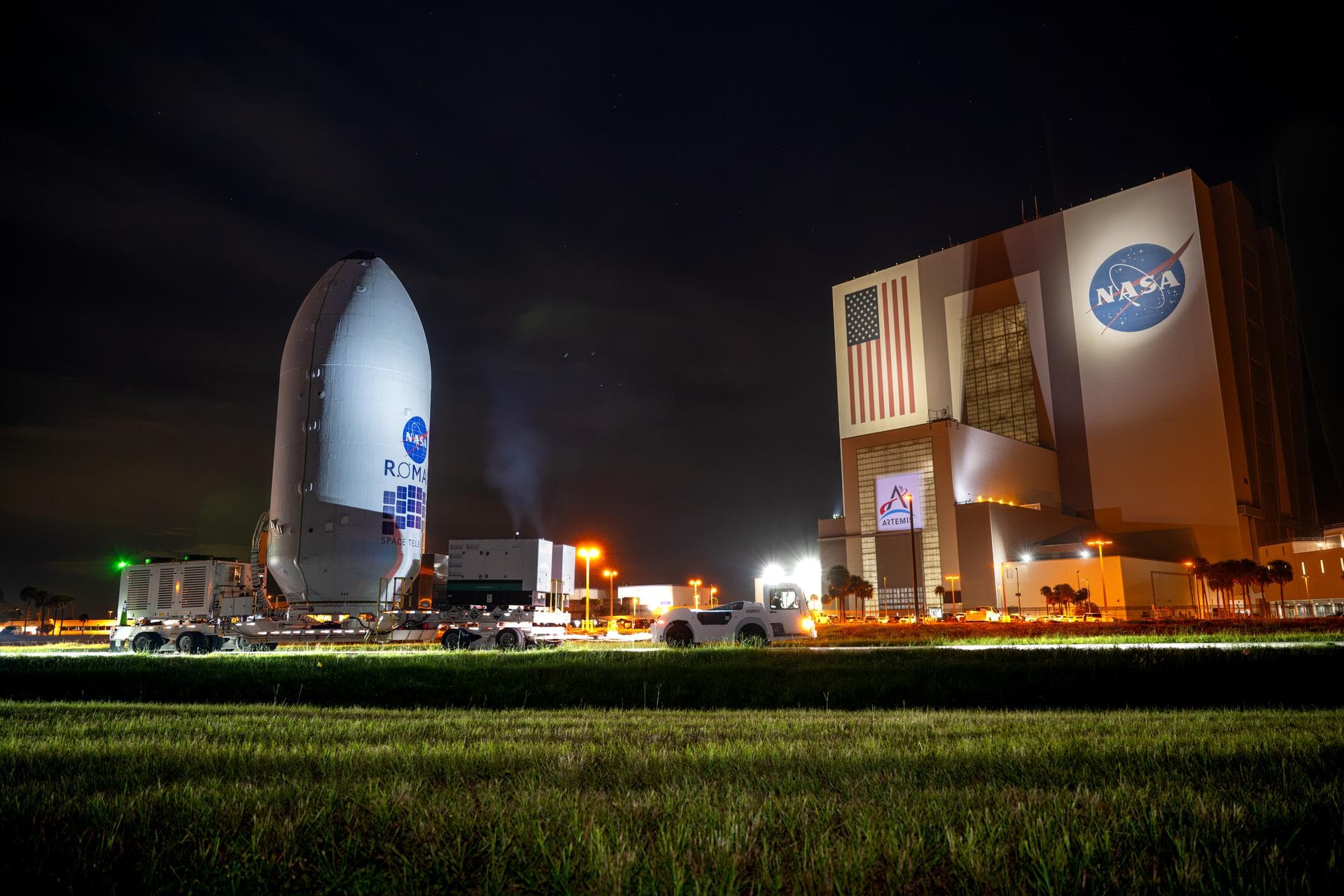The encapsulated Nancy Grace Roman Space Telescope on a transporter at Kennedy Space Center at night, with the Vehicle Assembly Building in the background
