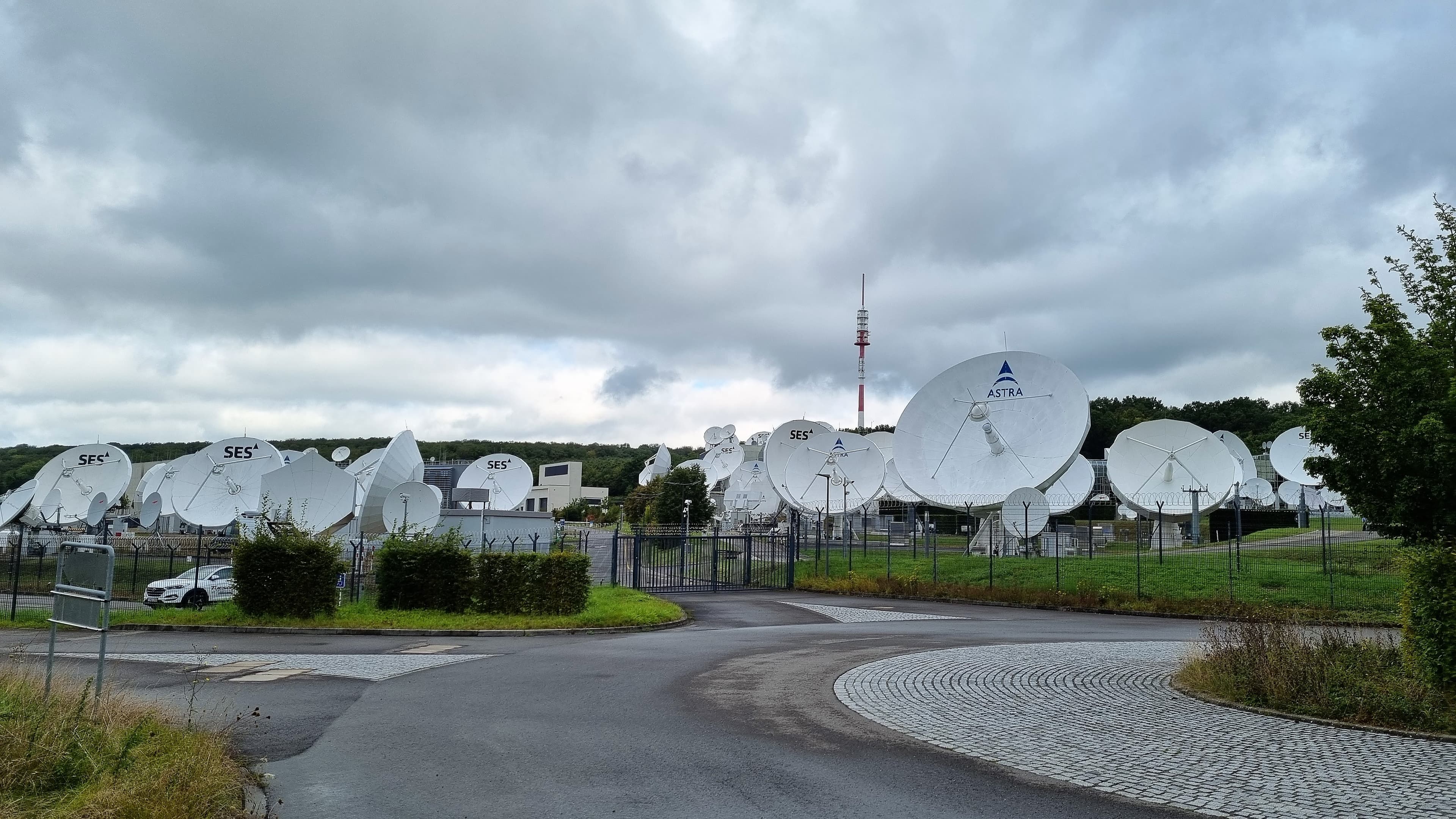 White SES and Astra satellite dishes behind a fence under cloudy skies in Betzdorf