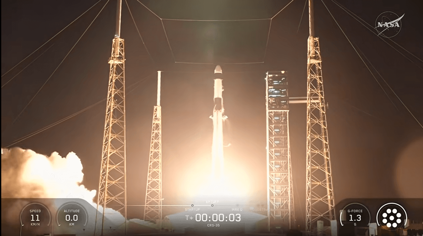 Falcon 9 rises between launch-pad towers above bright exhaust in a NASA broadcast frame