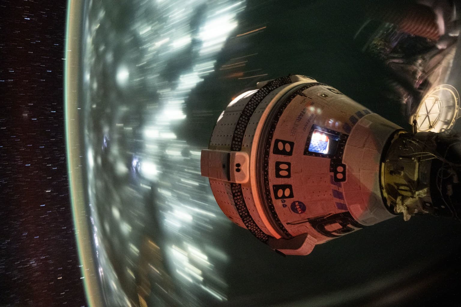 Starliner docked to the space station, with streaked city lights and stars behind the capsule
