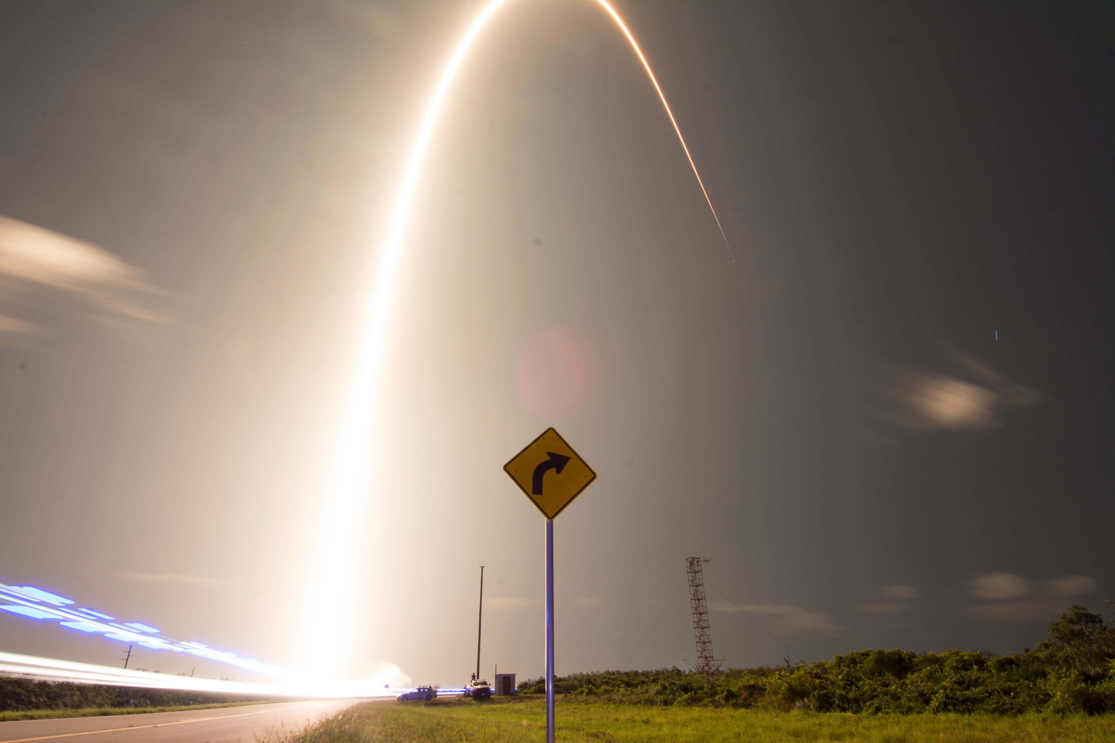 Long-exposure rocket trail curves over a road, directional sign and brightly illuminated Cape Canaveral launch site at night