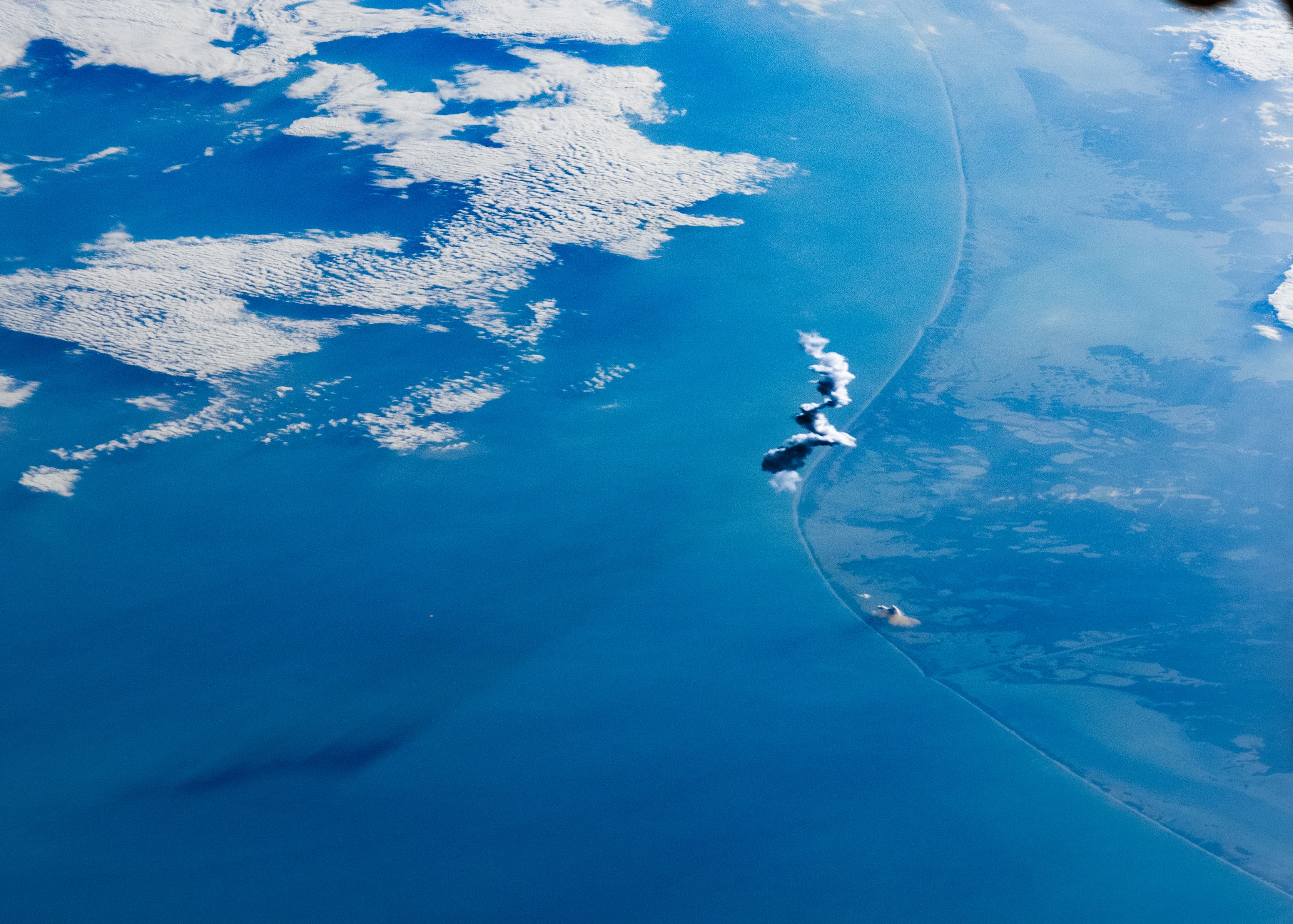 A white rocket plume casts a shadow over blue water beside the Texas coast, seen from orbit