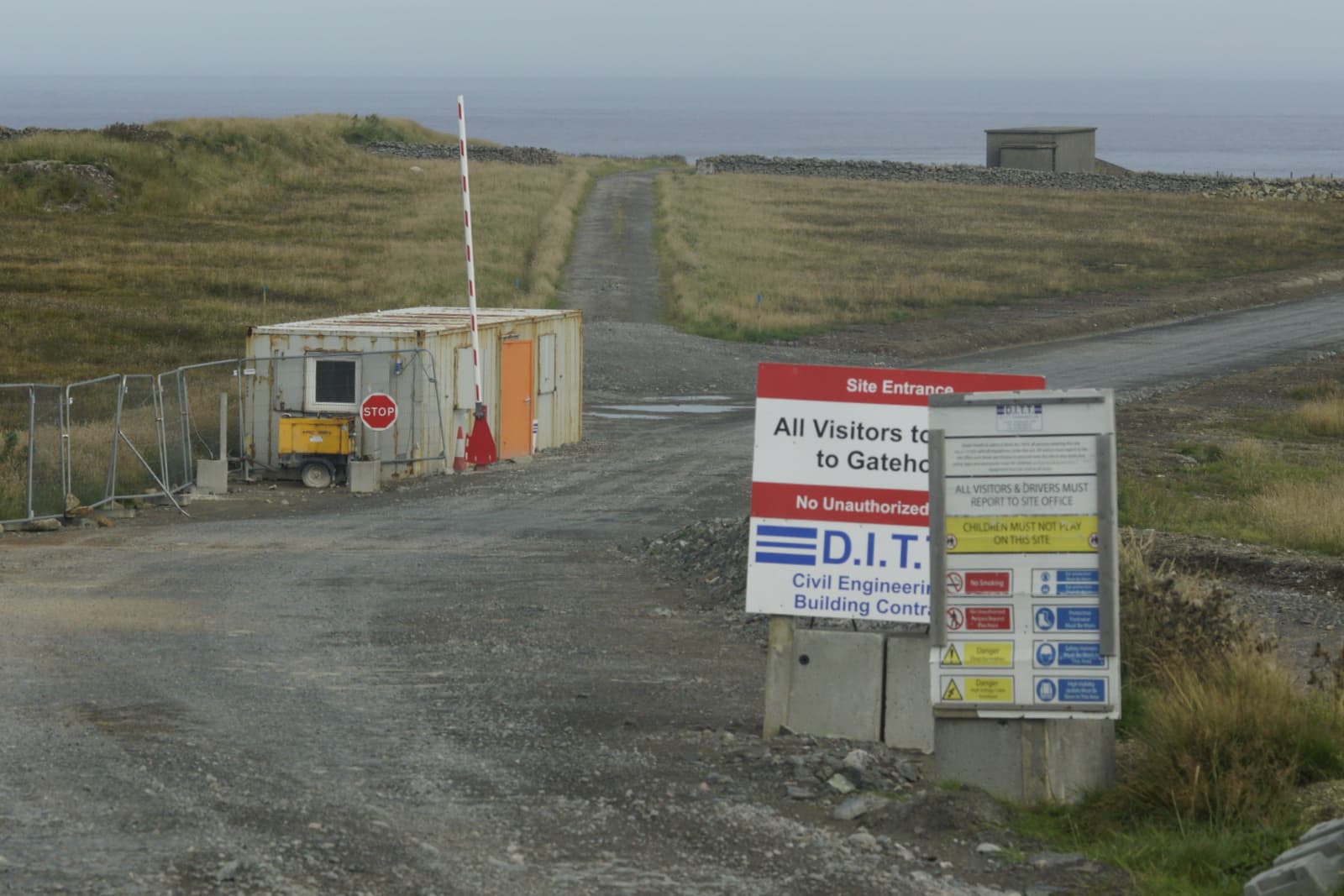 A gravel road passes a gatehouse and construction-site notices beside coastal grassland at SaxaVord